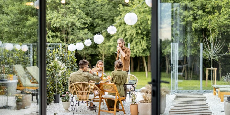 Friends having a festive dinner, gathering together at backyard of the house in nature. View through the window from the inside